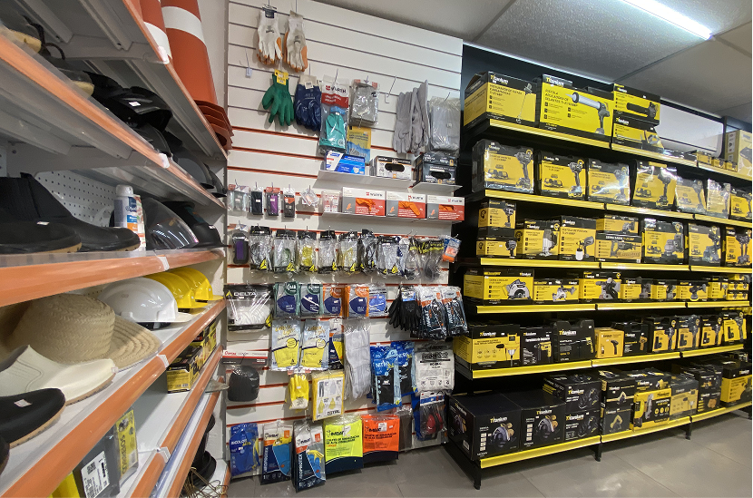Seção de ferramentas elétricas e Equipamentos de Proteção Individual (EPIs). À esquerda, prateleiras exibem calçados de segurança, luvas suspensas e capacetes de proteção nas cores branco e amarelo. À direita, um grande painel expositor preto exibe dezenas de caixas amarelas de ferramentas elétricas da linha profissional.